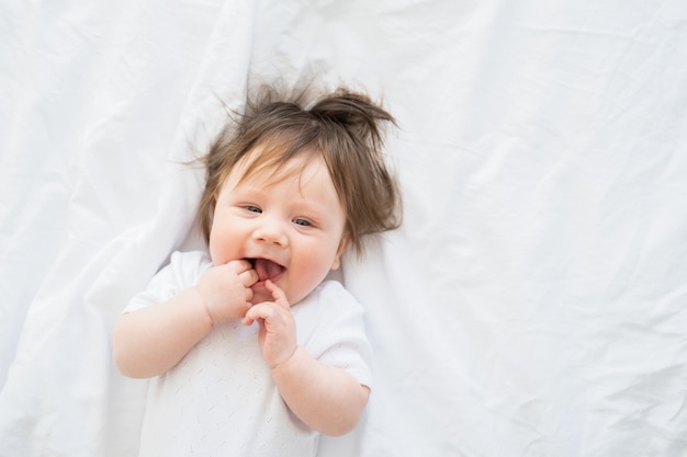 Porträt eines lustigen Babys mit dem Finger im Mund, das zu Hause auf einer weißen Bettwäsche lächelt und liegt.