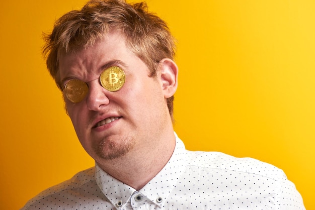 Porträt eines lustigen dicken Mannes mit finsterem Gesicht, Bitcoins in den Augen auf gelbem Hintergrund. Konzept von virtuellem digitalem Geld, Gier, Eitelkeit, Reichtum und Geiz