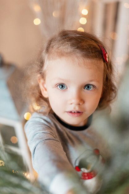 Porträt eines lustigen kleinen Mädchens mit schönen blauen Augen in modischen Pyjamas, die zu Hause einen Weihnachtsbaum schmücken. Winterurlaub und Wunder