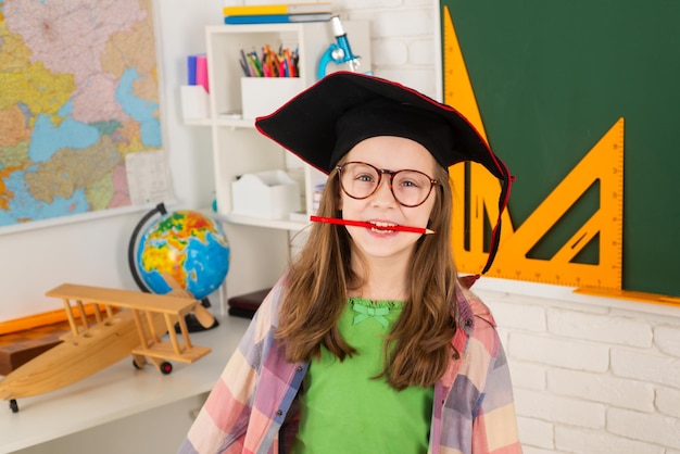 Porträt eines lustigen Schulmädchens mit Stift im Mund im Abschlusshut im Klassenzimmerkind, das studiert