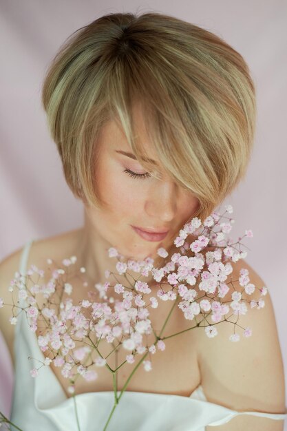 Porträt eines Mädchens auf einem rosa Hintergrund mit Blumen Zärtlichkeit und Weiblichkeit Die Sphäre der Schönheit und der Gesundheit der Frauen Kurzhaarschnitt blonde Haare