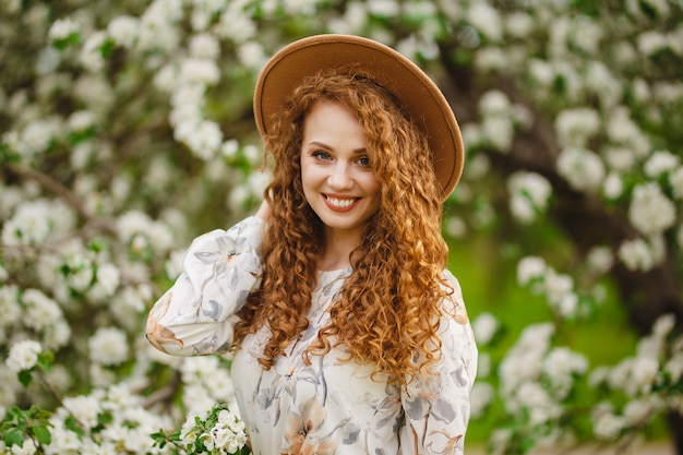 Porträt eines Mädchens, das an einem hellen sonnigen Frühlingstag lächelt und glücklich ist. Junge Frau mit dem lockigen Haar, das weißes Kleid und beigen Hut trägt, fühlt sich zufrieden, wenn sie im blühenden Park geht. Frühlingskonzept.