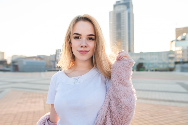 Porträt eines Mädchens im Teenageralter in rosa Kleidern und hellem Haar am Hintergrund der Sonne und der Stadtlandschaft