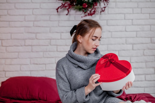 Porträt eines Mädchens in einem grauen Pullover mit einer herzförmigen roten Geschenkbox, das im Kopierraum des Valentinstagkonzepts in der Wohnung sitzt