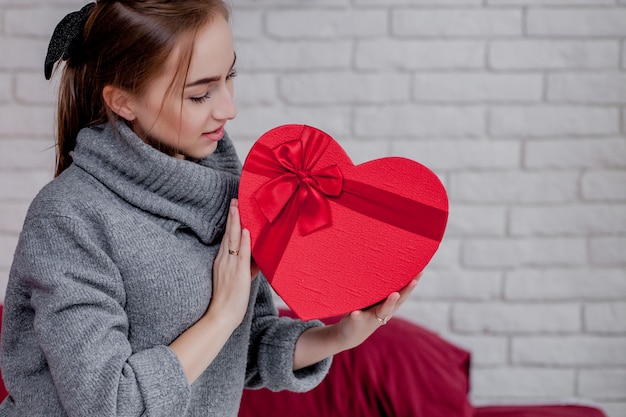 Porträt eines Mädchens in einem grauen Pullover mit einer herzförmigen roten Geschenkbox, die in der Wohnung sitzt, Valentinstagkonzept, Kopienraum