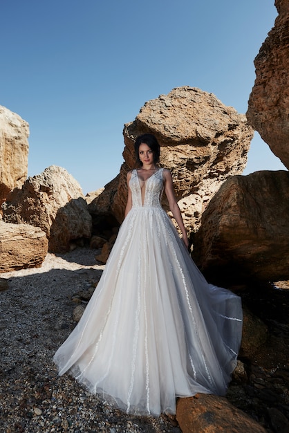 Porträt eines Mädchens in einem herrlichen Hochzeitskleid, das einen Fotografen auf dem Strand aufwirft. Die Braut ist auf den Felsen