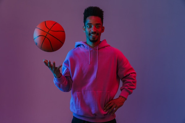 Porträt eines männlichen afroamerikanischen Mannes in einem bunten Hoodie, der mit Basketball isoliert über einer violetten Wand posiert