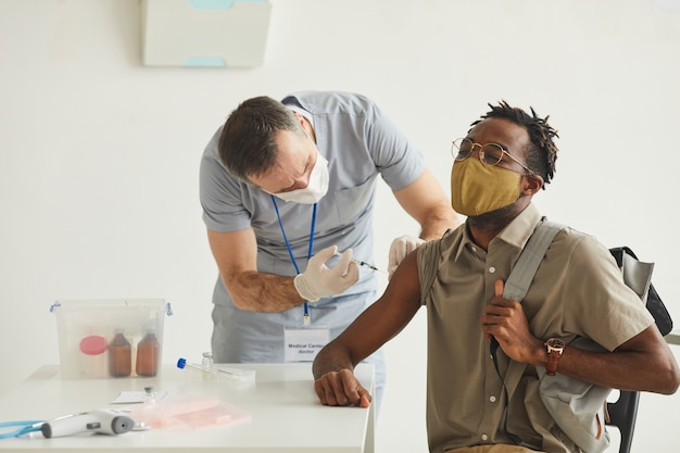 Porträt eines männlichen arztes, der jungen afroamerikanischen mann mit maske in einer medizinischen klinik impft, kopierraum