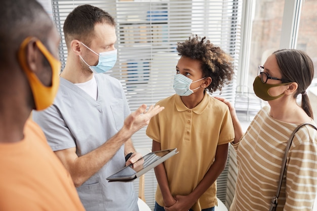 Porträt eines männlichen Arztes, der mit der Familie spricht, während er im Wartezimmer des Krankenhauses steht, alle tragen Masken