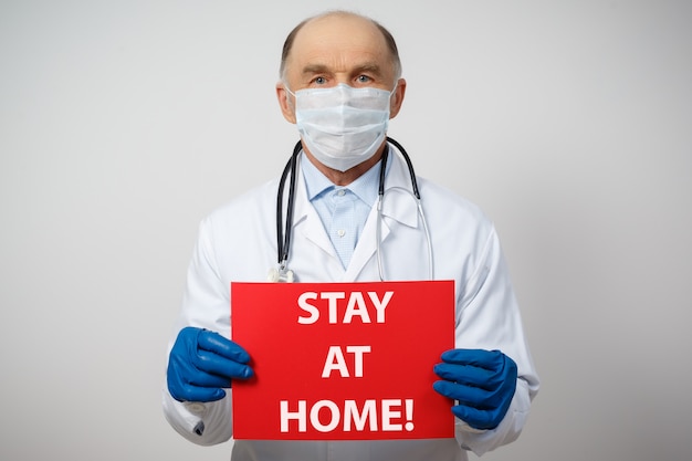 Porträt eines männlichen Arztes in einer medizinischen Schutzmaske und Handschuhen mit einem zu Hause bleibenden Plakat.