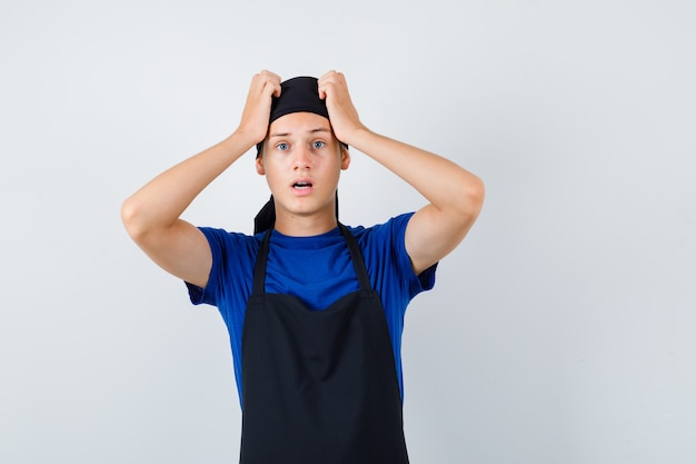 Porträt eines männlichen Teenagerkochs, der die Hände in T-Shirt, Schürze und schockierter Vorderansicht auf dem Kopf hält
