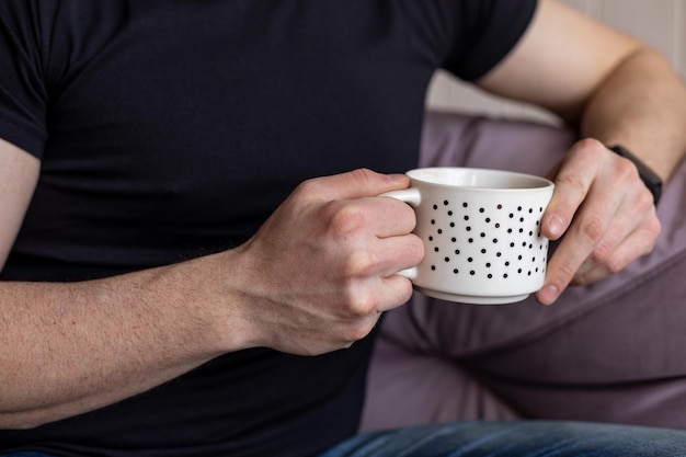 Porträt eines Mannes, der eine weiße Tasse Kaffee hält und auf einem weichen violetten Sofa in Freizeitkleidung sitzt