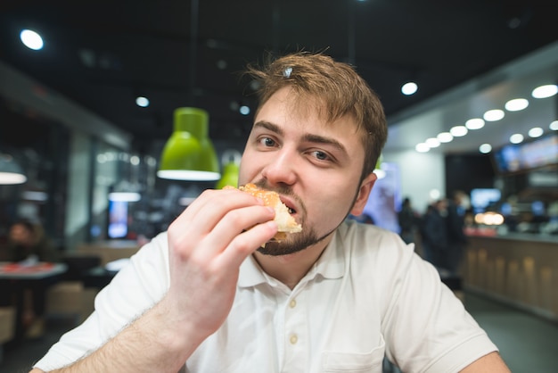 Porträt eines Mannes, der einen leckeren Burger beißt und in die Kamera schaut. Schnellimbiss im Fastfood-Restaurant.