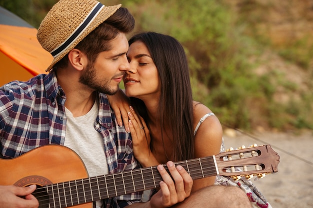 Porträt eines Mannes, der Gitarre für seine Freundin spielt, die im Campingzelt am Strand sitzt