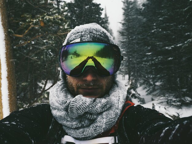 Porträt eines Mannes, der im Winter gegen Bäume steht und eine Skibrille mit Reflexion trägt