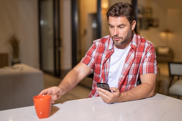 Porträt eines Mannes, der im Wohnzimmer auf sein Smartphone schaut. Mann redet, hält eine Tasse Kaffee und unterhält sich auf dem Sofa