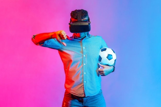 Porträt eines Mannes im Hemd, der eine Virtual-Reality-Brille trägt, mit Fußball steht, den Daumen nach unten zeigt, kein Fußballspiel mag. Studioaufnahme im Innenbereich isoliert auf buntem Neonlichthintergrund.