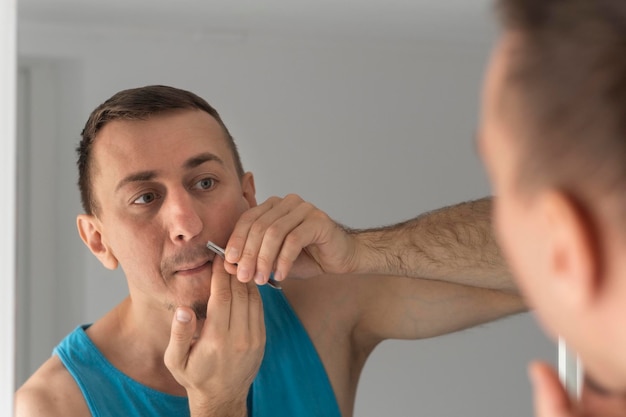 Porträt eines Mannes in blauem T-Shirt, der Haare mit einer Pinzette über seiner Oberlippe oder von der Nase zupft Spiegelung im Badezimmerspiegel