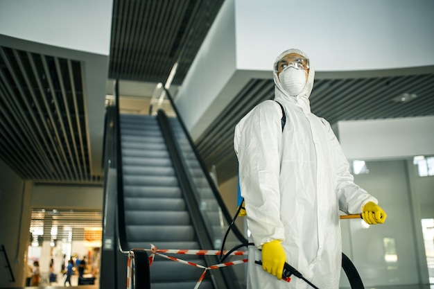 Porträt eines Mannes in einem Desinfektionsanzug, der Spray in der Nähe der Rolltreppe in einem leeren Einkaufszentrum hält. Ein Freiwilliger, der die öffentlichen Plätze aufräumt, um Covid-19 zu verhindern. Gesundheitsbewusstsein Konzept.