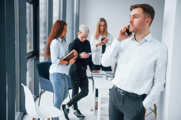 Porträt eines Mannes in formeller Kleidung und mit Telefon, das vor einem jungen erfolgreichen Team steht, das drinnen im Büro zusammenarbeitet und kommuniziert