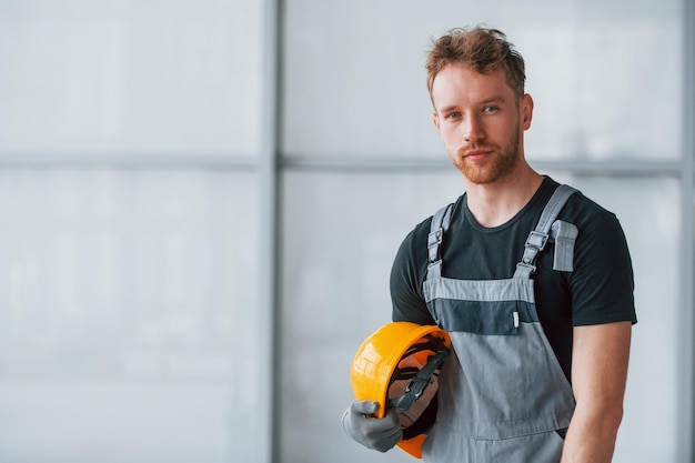 Porträt eines Mannes in grauer Uniform und mit orangefarbenem Schutzhelm in den Händen, der tagsüber in einem modernen großen Büro drinnen steht