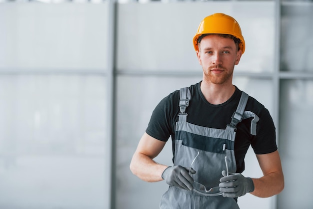 Porträt eines Mannes in grauer Uniform und orangefarbenem Schutzhelm, der tagsüber in einem modernen großen Büro drinnen steht