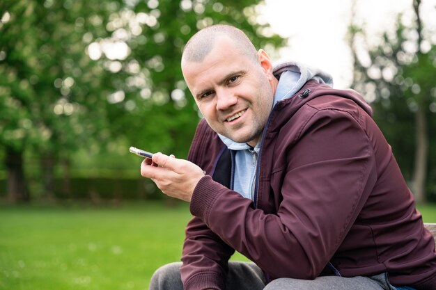 Porträt eines mannes von vierzig jahren, der in einem park sitzt und smartphone hält, in die kamera schaut und lächelt, jacke trägt.