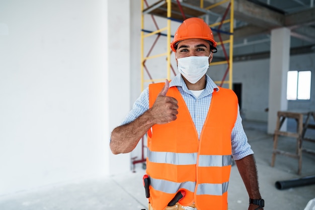 Porträt eines Mischlingsbauers in Arbeitskleidung und Bauarbeiterhelm mit medizinischer Maske aus nächster Nähe