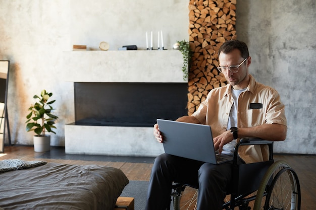 Porträt eines modernen erwachsenen Mannes im Rollstuhl mit Laptop zu Hause im Designer-Interieur, Kopierraum
