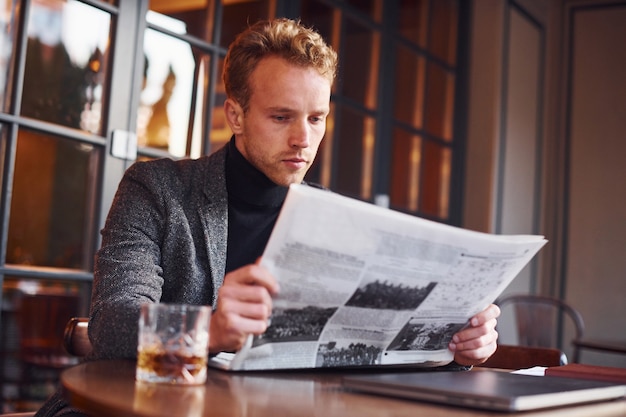 Porträt eines modernen jungen Mannes in formeller Kleidung, der im Café sitzt und Zeitung liest.