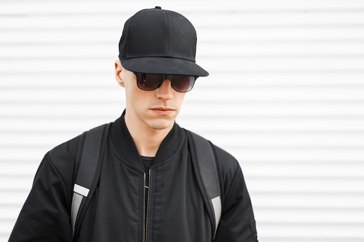 Foto porträt eines modischen mannes hipster in einer schwarzen mütze in stilvoller schwarzer sonnenbrille in einer trendigen jacke mit einem schwarzen rucksack auf seinen schultern an einer weißen wand. ernsthafter stilvoller amerikaner.