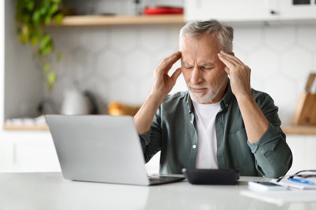 Porträt eines müden älteren Mannes mit Kopfschmerzen, der zu Hause am Laptop arbeitet
