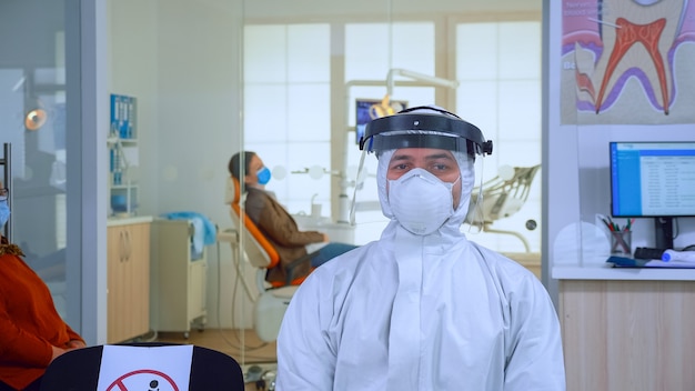 Porträt eines müden Arztes in der Zahnarztpraxis, der auf die Kamera schaut und einen Overall trägt und einen Gesichtsschutz auf einem Stuhl in der Wartezimmerklinik sitzt Konzept des neuen normalen Zahnarztbesuchs bei Ausbruch des Coronavirus.