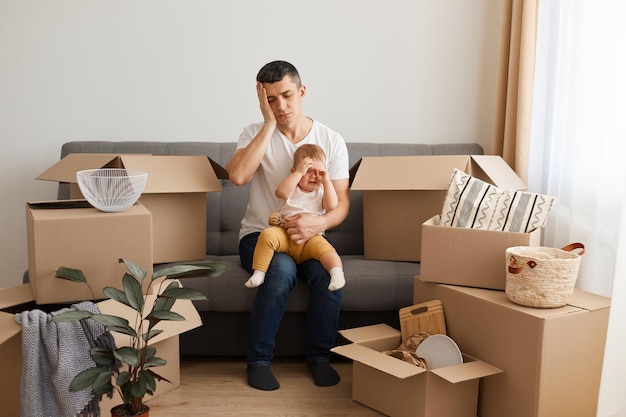 Porträt eines müden, erschöpften Mannes mit weißem T-Shirt und Jeans, der mit seiner kleinen Tochter auf dem Sofa sitzt, in ein neues Zuhause zieht, mit weinendem Kind posiert, unter schrecklichen Kopfschmerzen leidet, die Augen geschlossen hält