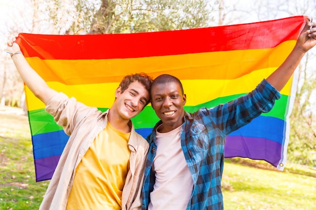 Porträt eines multiethnischen schwulen männlichen Paares, das bei Sonnenuntergang eine Regenbogen-lgbt-Symbolflagge hält