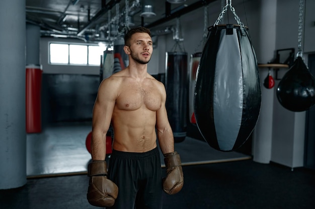 Porträt eines muskulösen männlichen Boxers, der neben einer schweren Tasche über dem Hintergrund der Boxhalle steht