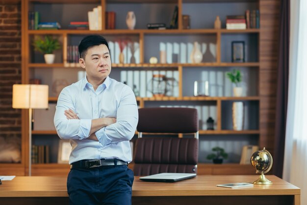 Porträt eines nachdenklichen Geschäftsmannes, ein Mann mit verschränkten Armen in einem Klassenzimmer, der arbeitet und aus dem Fenster auf der Rückseite schaut. Asiatisch denkt