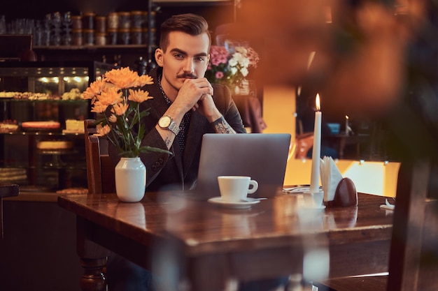 Porträt eines nachdenklichen gutaussehenden Geschäftsmannes mit stylischem Bart und Haaren in einem schwarzen Anzug, der mit der Hand am Kinn in einem Café mit offenem Laptop sitzt und in die Kamera schaut.