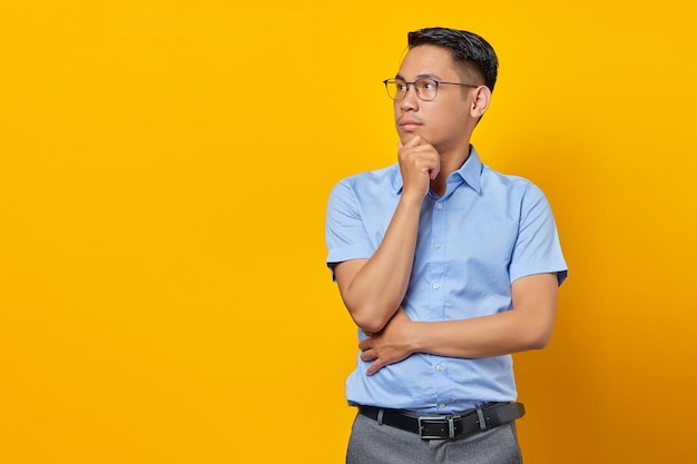 Porträt eines nachdenklichen jungen asiatischen Mannes mit Brille hält die Hand unter einem Kinn isoliert auf gelbem Hintergrund Geschäftsmann- und Unternehmerkonzept