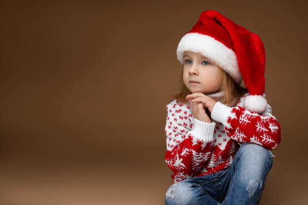 Porträt eines nachdenklichen kleinen Mädchens mit Weihnachtsmütze, rotem und weißem Strickpullover und Jeans, die Händchen am Kinn halten und wegschauen. Platz kopieren.