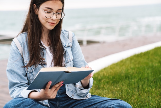 Porträt eines nachdenklichen, reizenden Teenager-Mädchens, das eine Brille trägt und ein Buch liest, während es auf grünem Gras am Meer sitzt