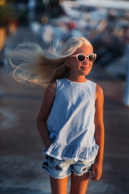 Porträt eines netten lächelnden zehnjährigen Mädchens mit Brille. Ein Mädchen in kurzen Hosen und ein blaues T-Shirt bei Sonnenuntergang nahe dem Meer. Türkei.