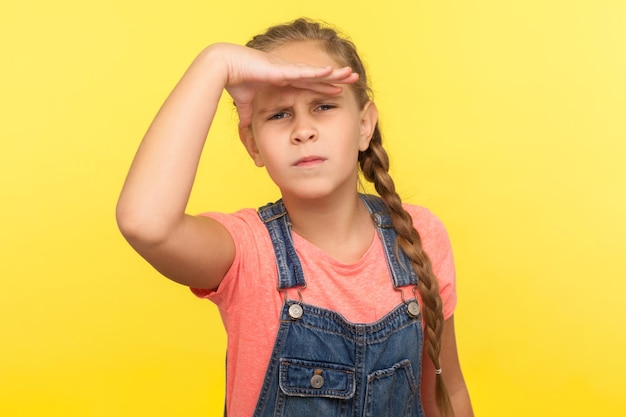 Porträt eines neugierigen, aufmerksamen, ernsten kleinen Mädchens in Denim-Overalls, das die Hand über die Augen hält und weit weg schaut, während es aus der Ferne beobachtet, wie man den Horizont beobachtet, Studioaufnahme isoliert auf gelbem Hintergrund