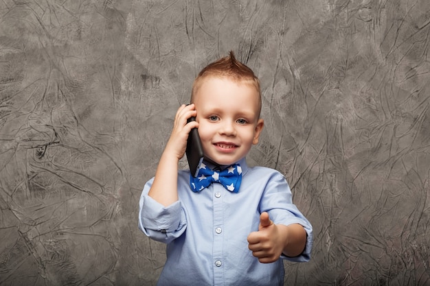 Porträt eines niedlichen kleinen Jungen im blauen Hemd und in der Fliege mit Handy gegen graue Textur