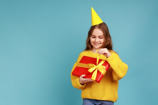 Porträt eines niedlichen kleinen Mädchens im Partykegel, das eine Geschenkbox öffnet, die positive glückliche Emotionen ausdrückt, die einen gelben Pullover im lässigen Stil tragen Studioaufnahme isoliert auf blauem Hintergrund