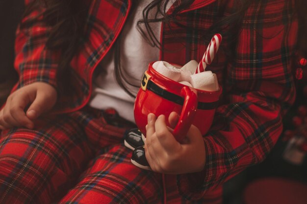 Porträt eines offenen asiatischen lächelnden kleinen Mädchens mit rotem Weihnachtsbecher mit Marshmallows und Zuckerstange