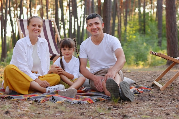 Porträt eines optimistischen, zufriedenen jungen Paares, das sich zusammen mit seinen Töchtern im Wald ausruht, die lässige Kleidung tragen, auf dem Boden sitzen und lächelnd in die Kamera schauen