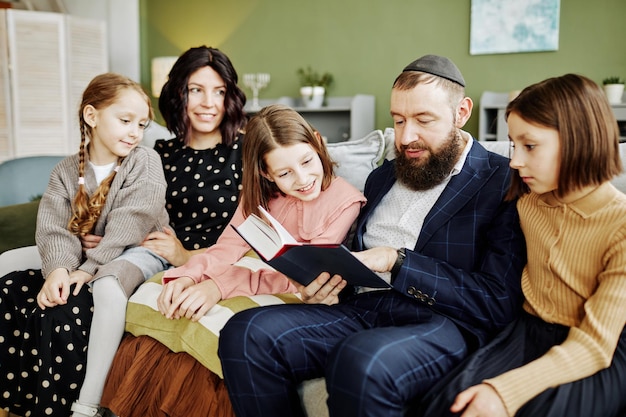 Porträt eines orthodoxen jüdischen Mannes, der Kippa trägt, während er vielen Kindern zu Hause ein Buch vorliest