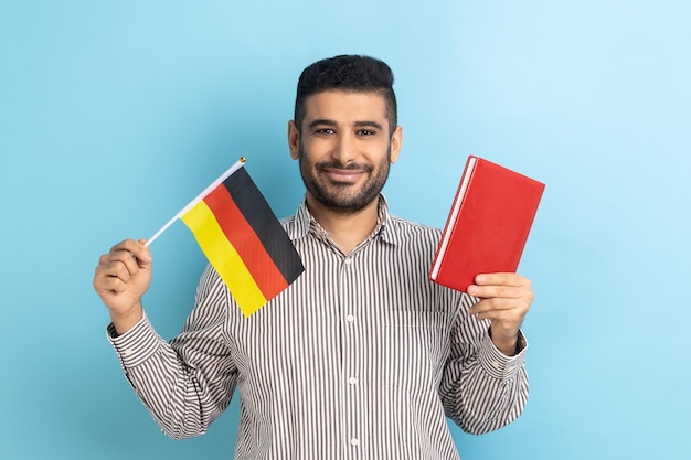 Porträt eines positiv lächelnden Mannes mit Buch und deutscher Flagge, der davon träumt, in Deutschland zu studieren, Bildung im Ausland mit gestreiftem Hemd. Studioaufnahme im Innenbereich isoliert auf blauem Hintergrund