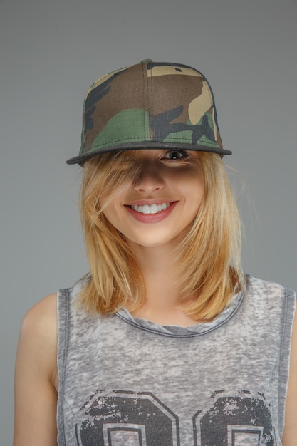 Porträt eines positiven blonden Mädchens in Militärmütze isoliert auf grauem Hintergrund.